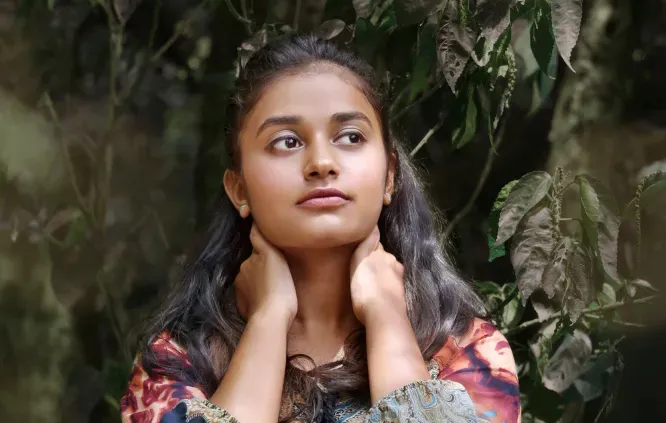 Woman with long hair posing with hands on neck, standing amidst green foliage in natural light.